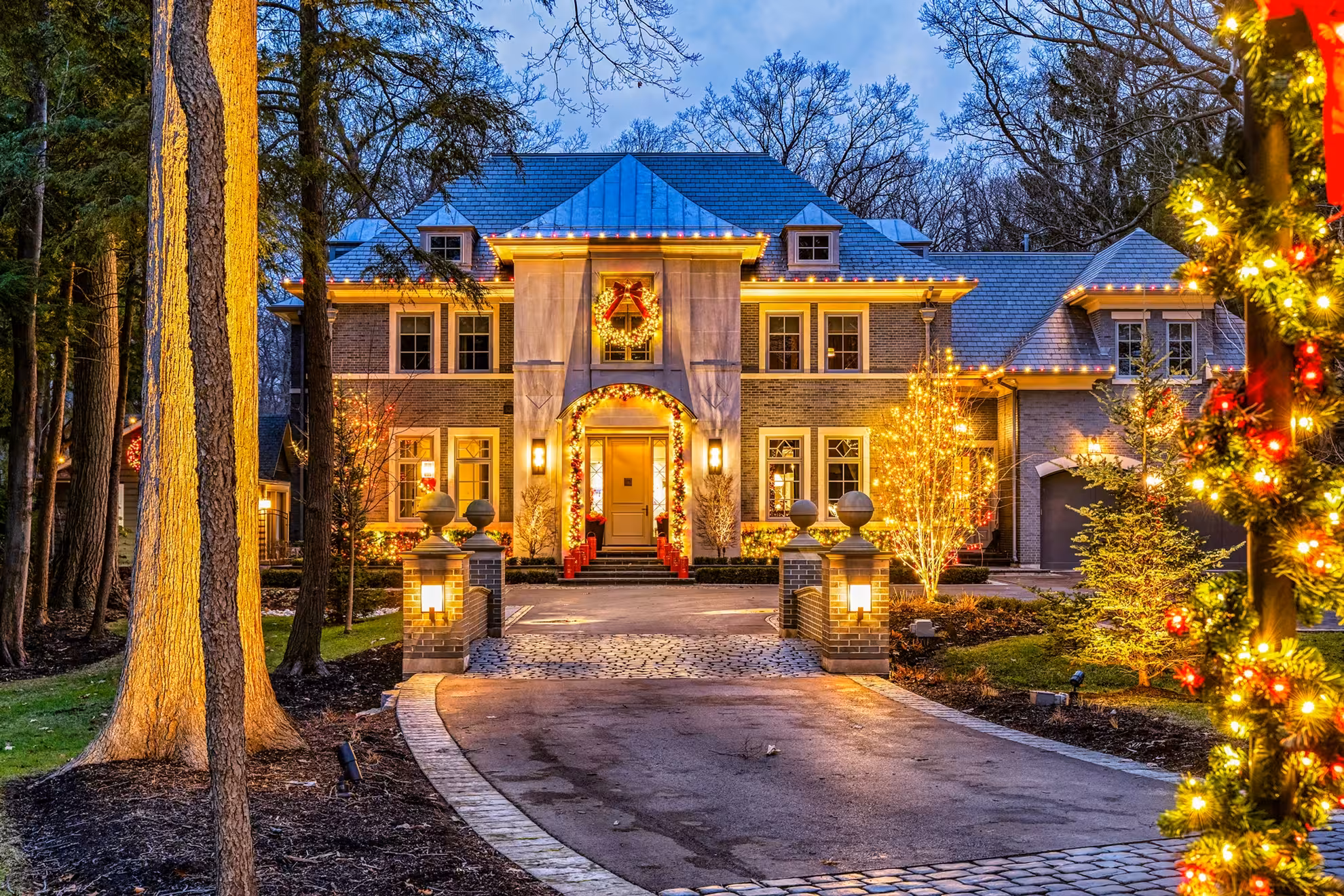 A Massachusetts estate home beautifully lit for Christmas at dusk — roofline lights, wreaths, garland and wrapped trees